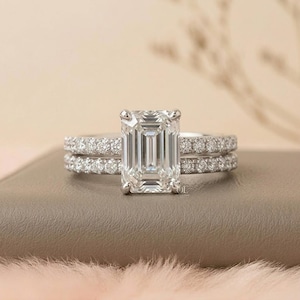 May include: A silver engagement ring set featuring an emerald-cut center stone and a pav&eacute; band. The set includes a matching band with small, round accent stones. The rings are displayed on a neutral surface with a soft, blurred background.