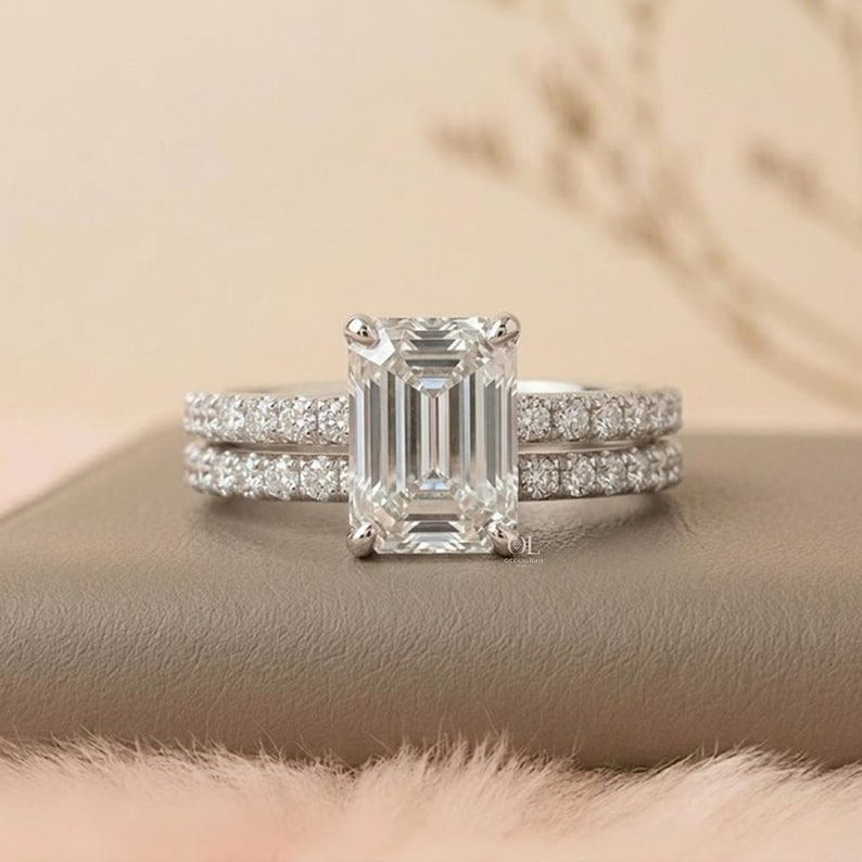 May include: A silver engagement ring set featuring an emerald-cut center stone and a pav&eacute; band. The set includes a matching band with small, round accent stones. The rings are displayed on a neutral surface with a soft, blurred background.