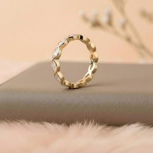May include: A gold ring with a repeating pattern of marquise-cut gemstones. The ring is set on a neutral-toned surface, with a soft, blurred background. The ring's design features alternating gold and clear gemstone elements.