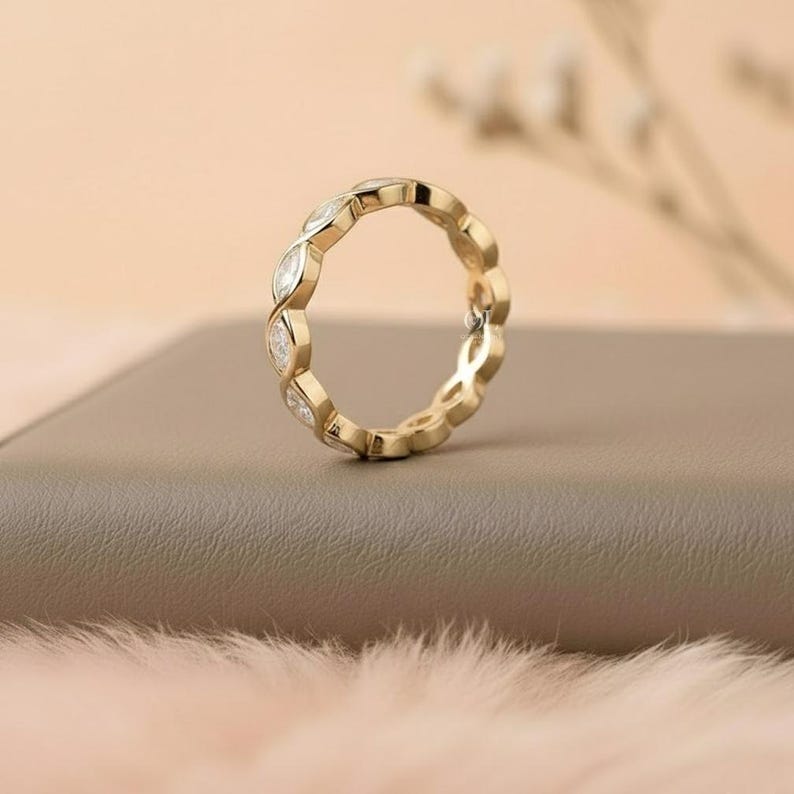 May include: A gold ring with a repeating pattern of marquise-cut gemstones. The ring is set on a neutral-toned surface, with a soft, blurred background. The ring's design features alternating gold and clear gemstone elements.