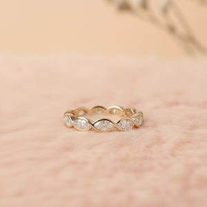 May include: A gold band ring with marquise-cut gemstones set in a continuous pattern. The ring is displayed on a pink surface, showcasing its intricate design and sparkle. The ring is a piece of jewelry.