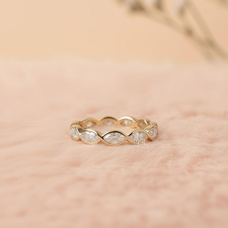 May include: A gold band ring with marquise-cut gemstones set in a continuous pattern. The ring is displayed on a pink surface, showcasing its intricate design and sparkle. The ring is a piece of jewelry.