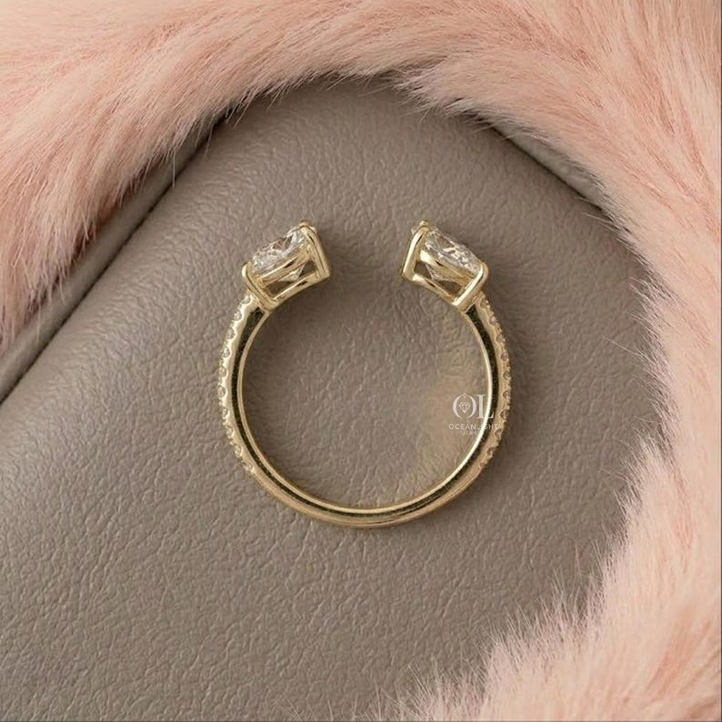 May include: A gold ring with two pear-shaped gemstones set at opposite ends. The band is encrusted with small, sparkling stones. The ring is set against a gray surface with a pink, fluffy background.