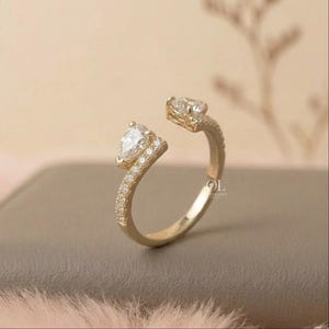 May include: A gold-colored open ring featuring two pear-shaped, clear gemstones. The ring's band is adorned with small, sparkling stones. The design is elegant and modern, suitable for various occasions.