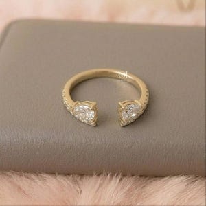 May include: A gold-colored open ring featuring two pear-shaped, clear gemstones. The ring band is encrusted with small, clear stones. The ring is set against a neutral background.