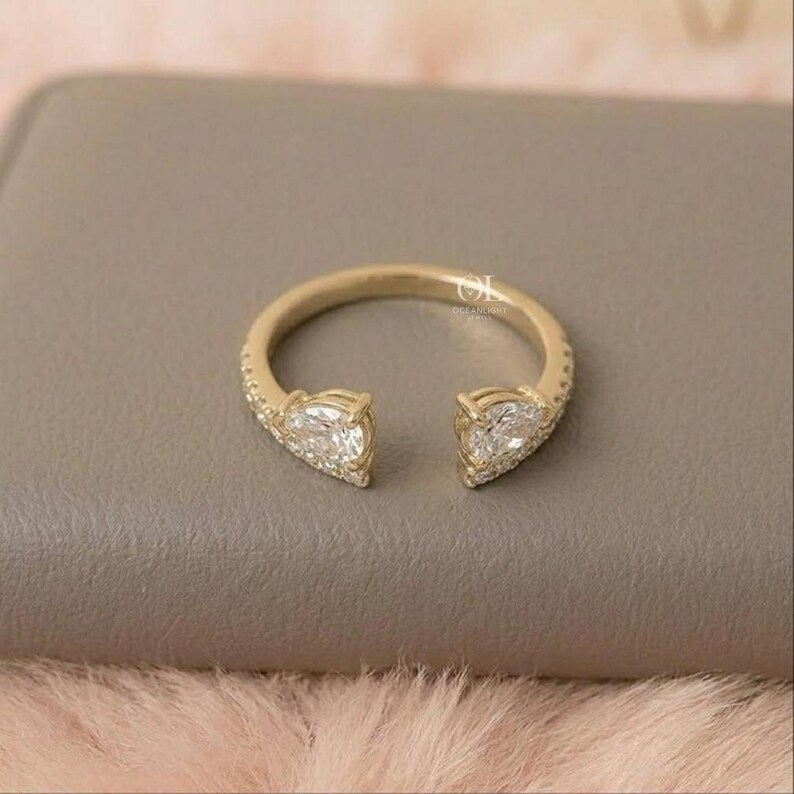 May include: A gold-colored open ring featuring two pear-shaped, clear gemstones. The ring band is encrusted with small, clear stones. The ring is set against a neutral background.