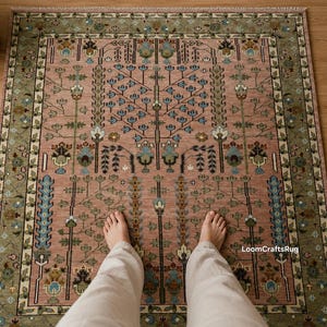May include: A rectangular area rug featuring a dusty pink field with a central tree of life motif in blue, green, and cream. The border is a muted olive green with floral patterns. The text LoomCraftsRug is printed in the lower right corner.