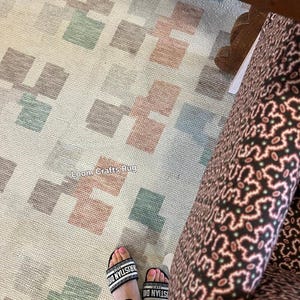 May include: A beige rug with a geometric pattern of squares in shades of brown, green, and pink. The text "Loom Crafts Rug" is visible on the rug. Black and white sandals with the text "Christian Dior" are also visible.