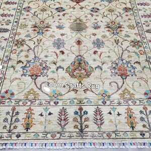 May include: Cream-colored rug with an intricate floral pattern in shades of blue, orange, and green. The rug features a detailed border and fringe along the edges. The text "Loom Crafts Rug" is visible on the rug.