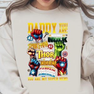May include: Cream-colored sweatshirt with a graphic featuring superhero fists and text. The text includes "Daddy You Are" and references to Spiderman, Hulk, Thor, Iron Man, and Captain America. The phrase "You Are My Super Hero" is also present.