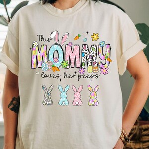 May include: A cream-colored t-shirt with a colorful Easter-themed graphic. The design includes the text "This MOMMY loves her peeps" with bunny ears, carrots, flowers, and decorated eggs. Below the text are four bunny silhouettes.