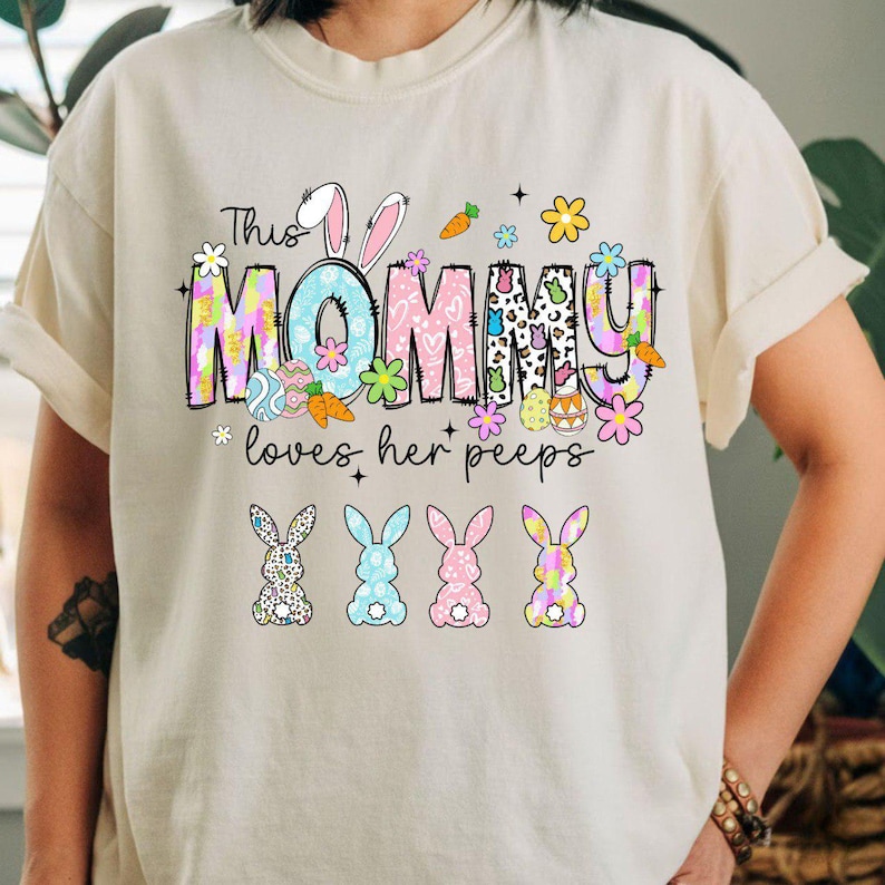 May include: A cream-colored t-shirt with a colorful Easter-themed graphic. The design includes the text "This MOMMY loves her peeps" with bunny ears, carrots, flowers, and decorated eggs. Below the text are four bunny silhouettes.