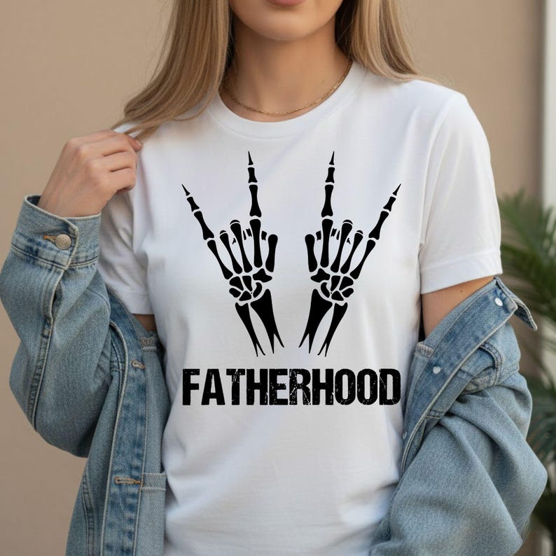 May include: White t-shirt featuring a black graphic of two skeleton hands making a rock and roll gesture. The word "FATHERHOOD" is printed in bold, black letters below the hands. The shirt is a casual style.