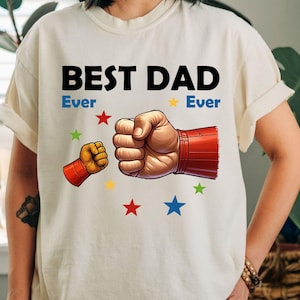 May include: Cream-colored t-shirt with the text "BEST DAD Ever Ever" in black and blue. The graphic features two fists, one large and one small, with red and yellow accents, surrounded by colorful stars.