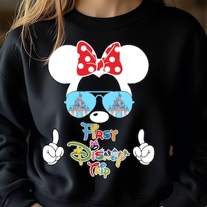 May include: Black sweatshirt featuring a Minnie Mouse design with a red polka dot bow, sunglasses reflecting a castle, and the text "First Disney Trip." White gloved hands point upwards on either side of the text.
