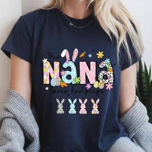 May include: Navy blue t-shirt with the text "Nana loves her peeps." The word "Nana" is decorated with Easter-themed graphics, including bunny ears, flowers, and eggs. Four colorful bunny illustrations are at the bottom.