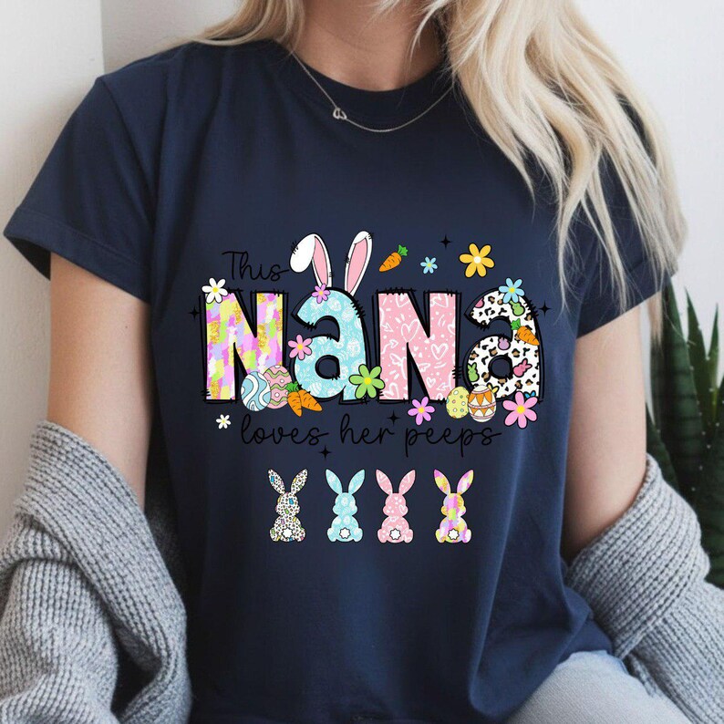 May include: Navy blue t-shirt with the text "Nana loves her peeps." The word "Nana" is decorated with Easter-themed graphics, including bunny ears, flowers, and eggs. Four colorful bunny illustrations are at the bottom.