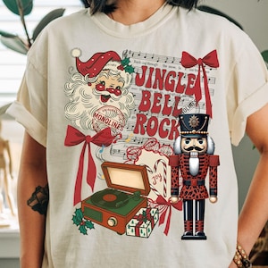 Puede incluir: Camiseta color crema con un diseño navideño festivo. El gráfico incluye a Papá Noel, un cascanueces, notas musicales y las palabras "Jingle Bell Rock". Elementos adicionales incluyen un tocadiscos, bastones de caramelo y una caja de regalo.