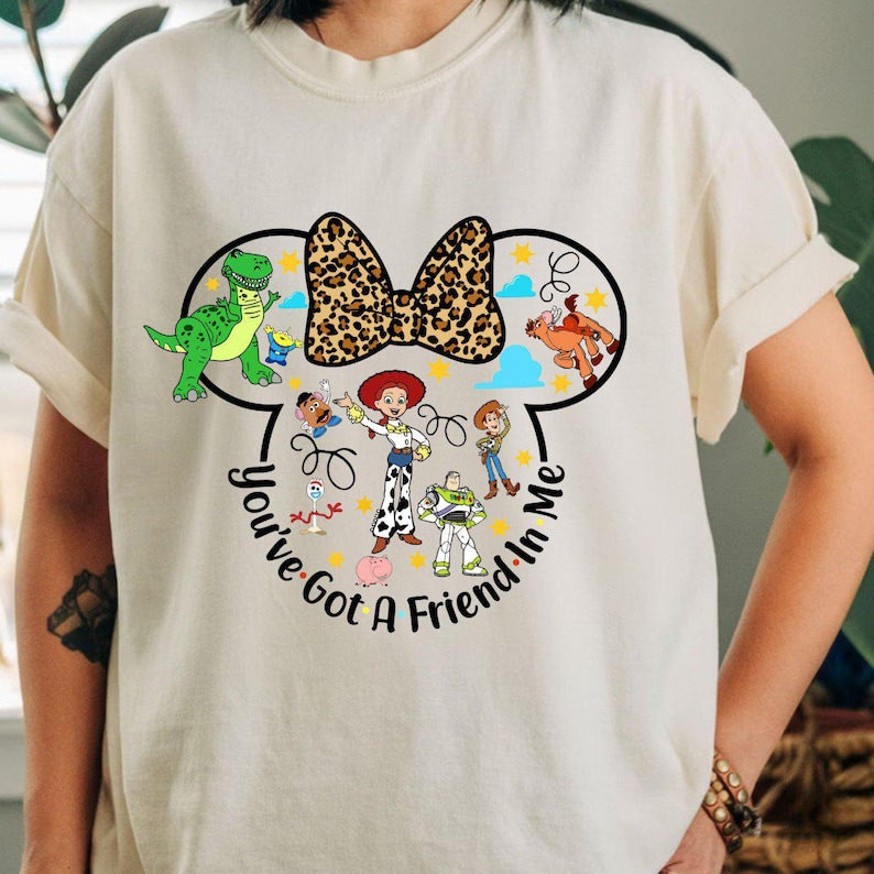 K&ouml;nnte beinhalten: Cremefarbenes T-Shirt mit einer Grafik von Disney's Toy Story-Charakteren in einer Mickey Mouse-Ohren-Umrisslinie. Das Design umfasst eine Leopardenmuster-Schleife, den Text "You've Got A Friend In Me" und Charakterillustrationen.