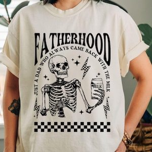 May include: Cream-colored t-shirt with a black graphic featuring a skeleton holding a milk carton. The text reads "FATHERHOOD" and "JUST A DAD WHO ALWAYS CAME BACK WITH THE MILK". Includes lightning bolts and a checkered pattern.