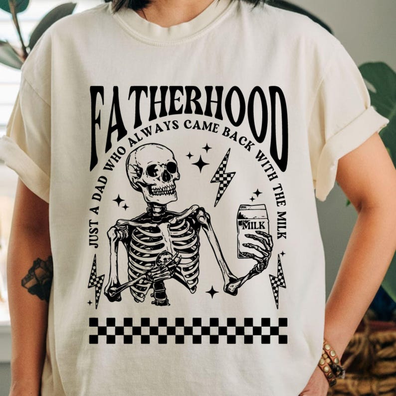 May include: Cream-colored t-shirt with a black graphic featuring a skeleton holding a milk carton. The text reads "FATHERHOOD" and "JUST A DAD WHO ALWAYS CAME BACK WITH THE MILK". Includes lightning bolts and a checkered pattern.