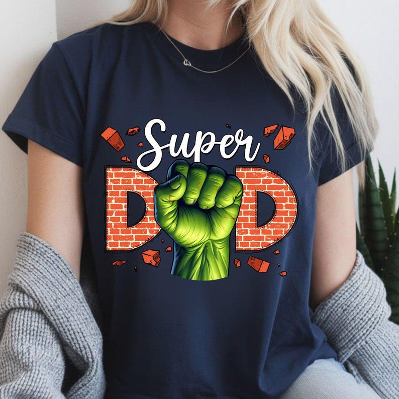 May include: Navy blue t-shirt with the words "Super Dad" in white script and brick-style letters. A green fist graphic is in the center. The shirt is a crew neck and short-sleeved.