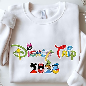 May include: White crewneck sweatshirt with a colorful "Disney Trip 2026" graphic. The text features various Disney character-themed designs, including Mickey Mouse, Minnie Mouse, and Donald Duck. The sweatshirt is made of a soft material.