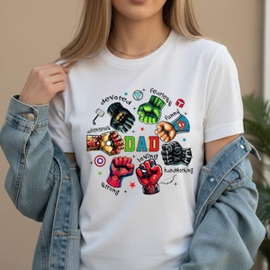 May include: White t-shirt with a colorful graphic featuring superhero fists and the word "DAD" in green. Surrounding the word are phrases like "devoted," "fearless," and "loving."