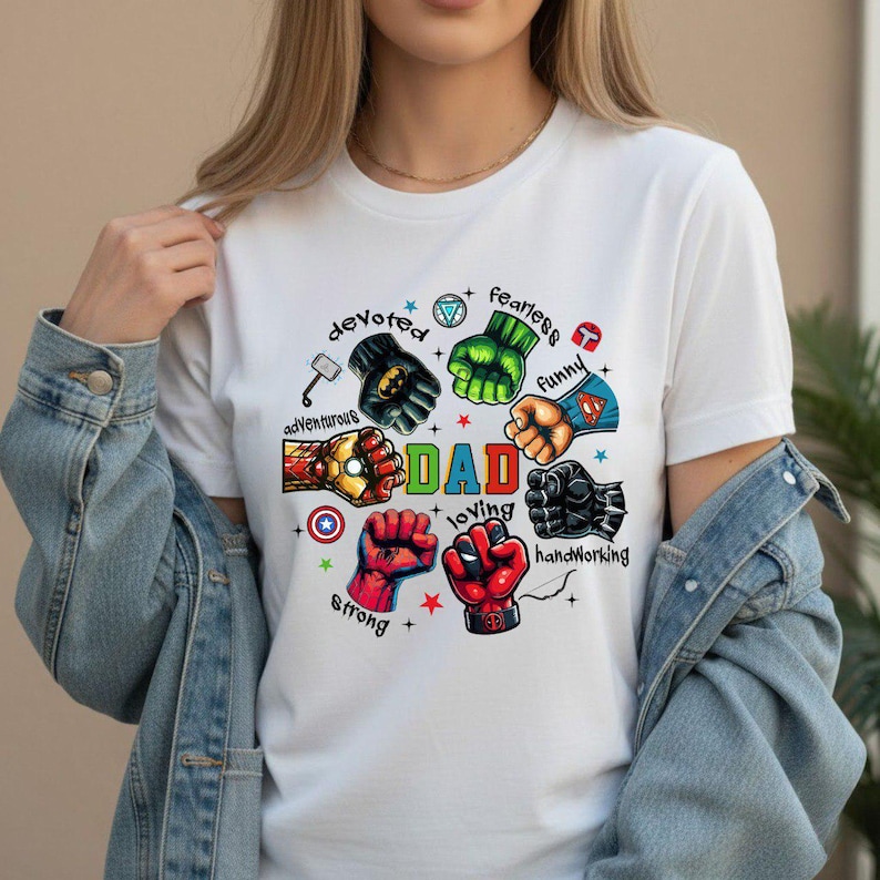 May include: White t-shirt with a colorful graphic featuring superhero fists and the word "DAD" in green. Surrounding the word are phrases like "devoted," "fearless," and "loving."