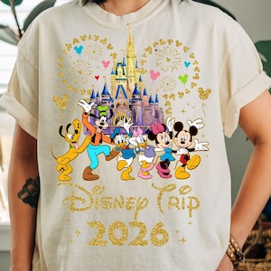 May include: Cream-colored t-shirt featuring a graphic of Disney characters, including Mickey and Minnie Mouse, in front of a castle. The words "Disney Trip 2026" are printed in gold glitter lettering, with fireworks and Mickey Mouse head shapes.