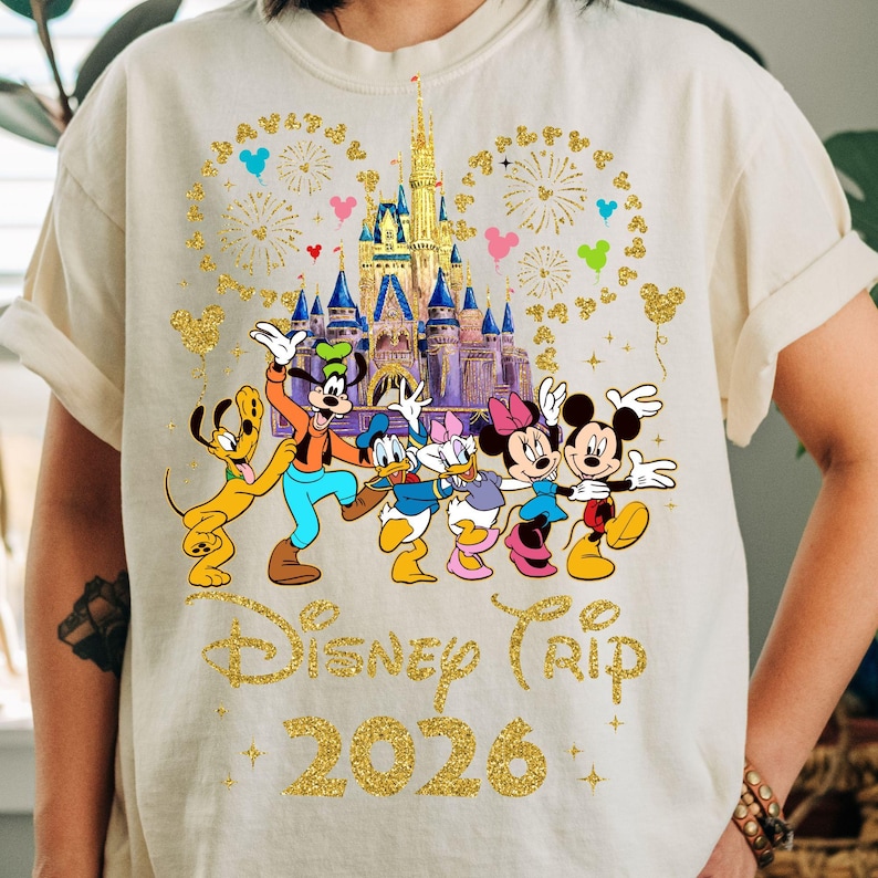 May include: Cream-colored t-shirt featuring a graphic of Disney characters, including Mickey and Minnie Mouse, in front of a castle. The words "Disney Trip 2026" are printed in gold glitter lettering, with fireworks and Mickey Mouse head shapes.