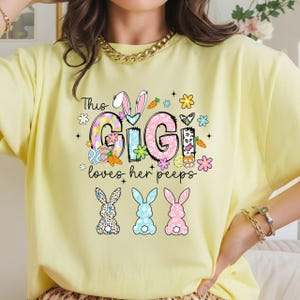 May include: A light yellow t-shirt with the text "This Gigi loves her peeps" in a colorful design. The design includes Easter eggs, carrots, flowers, and three bunny silhouettes.