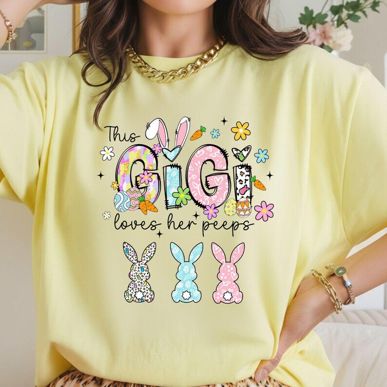 May include: A light yellow t-shirt with the text "This Gigi loves her peeps" in a colorful design. The design includes Easter eggs, carrots, flowers, and three bunny silhouettes.