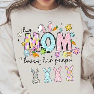 May include: Beige sweatshirt with a colorful Easter-themed graphic. The design includes the words "This MOM loves her peeps" with bunny ears, flowers, carrots, and Easter eggs. Four bunny silhouettes are at the bottom.