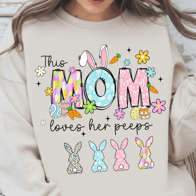 May include: Beige sweatshirt with a colorful Easter-themed graphic. The design includes the words "This MOM loves her peeps" with bunny ears, flowers, carrots, and Easter eggs. Four bunny silhouettes are at the bottom.