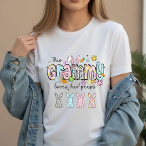 May include: White t-shirt with the text "This Grammy loves her peeps" in colorful, decorative lettering. The design includes Easter-themed graphics like bunnies, carrots, and flowers. Below the text are four bunny illustrations.