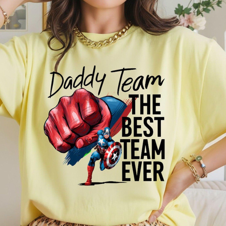 May include: A light yellow t-shirt with the text "Daddy Team" above a graphic of a red and blue fist and Captain America. The text "THE BEST TEAM EVER" is below the graphic.