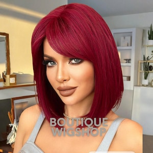 May include: A woman with a red bob wig with bangs. The wig is a vibrant shade of red. The text "BOUTIQUE WIGSHOP" is visible on the image. The woman is wearing a silver top and a gold necklace.