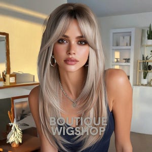 Puede incluir: Una mujer con una peluca rubia larga y en capas con flequillo. La peluca tiene raíces oscuras y reflejos rubios claros. El texto "BOUTIQUE WIGSHOP" es visible en la parte frontal de la imagen. Lleva un collar plateado y pendientes de aro.