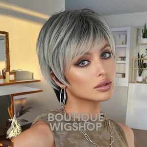 May include: A woman with a short, layered wig in shades of gray and blonde. The wig has a side-swept fringe and is styled to frame the face. She is wearing large silver hoop earrings and a silver chain necklace. The text "BOUTIQUE WIGSHOP" is visible.