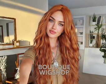 May include: A woman with long, wavy, auburn hair. She is wearing a beige top and multiple silver necklaces. The text "BOUTIQUE WIGSHOP" is overlaid on the image. The background includes a mirror, shelves, and plants.