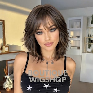 May include: A shoulder-length brown wig with layered ends and wispy bangs. The wig features a natural-looking part and subtle highlights. Overlaid on the image is the white text BOUTIQUE WIGSHOP. The wig is worn with a black top featuring white stars.
