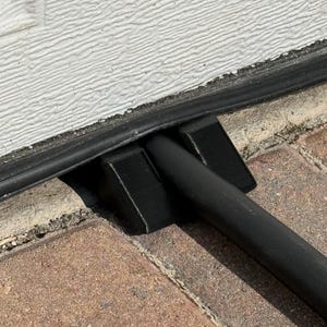 May include: Black metal bar with two black rectangular supports, resting on a brick surface. The bar is positioned near a white textured wall and a black rubber strip.