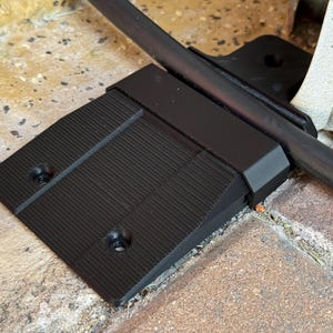 May include: A black plastic cable protector with a ridged surface and two screw holes. A black metal bar rests on top of the protector. The protector is on a brick surface.