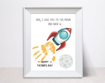 May include: A white framed print with a yellow handprint, a red and blue rocket, and a gray moon. The text reads "Dad, I love you to the moon and back" and "Happy Father's Day".
