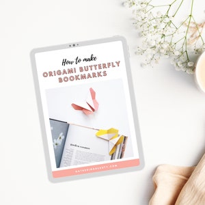 K&ouml;nnte beinhalten: Ein digitales Tablet zeigt Anweisungen zum Falten von Origami-Schmetterlings-Lesezeichen. Der Bildschirm zeigt einen rosa und wei&szlig;en Origami-Schmetterling und ein gelb-wei&szlig;es Origami-Lesezeichen. Der Text auf dem Bildschirm lautet "How to make ORIGAMI BUTTERFLY BOOKMARKS".