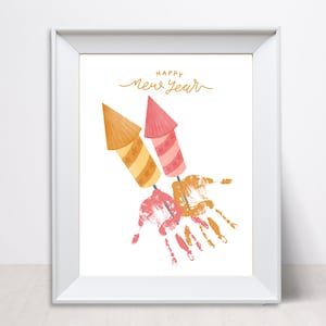May include: A white framed print with the text "Happy New Year" in gold lettering. The print features two handprint rockets with red, orange, and yellow paint.