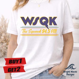 May include: White t-shirt featuring the text "WSQK The Squawk 94.5 FM" in purple, green, and yellow. The shirt has a crew neck and short sleeves. A red banner with "BUY 1 GET 2" is visible.
