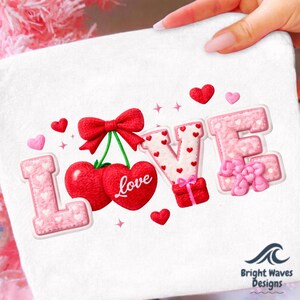 May include: White fabric featuring the word "LOVE" in a decorative design. The "L" and "E" are pink with white hearts, the "V" is white with red hearts, and the "O" is represented by two red heart-shaped cherries with a red bow.