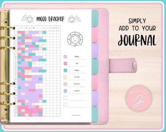 May include: A pink planner with a mood tracker chart and colour-coded sections for emotions. The chart is filled with pastel colours. Text on the page reads "MOOD TRACKER" and "SIMPLY ADD TO YOUR JOURNAL". A round sticker says "MADE TO J".